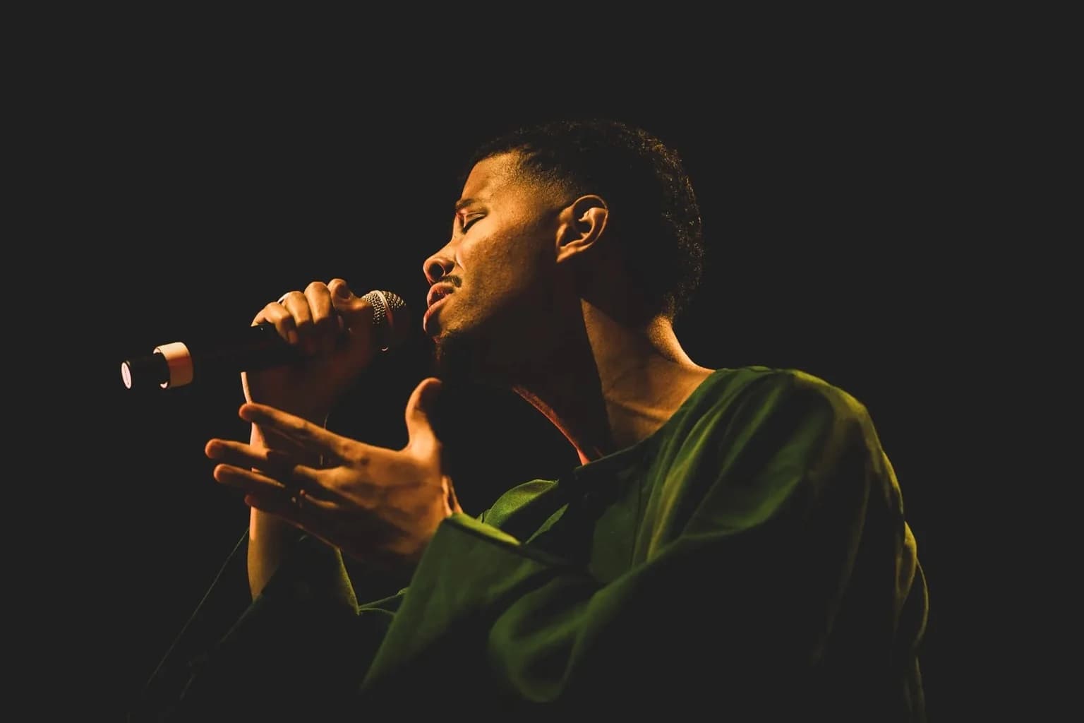 Mustafa sings onstage in a green shirt, holding a microphone against a dark background.