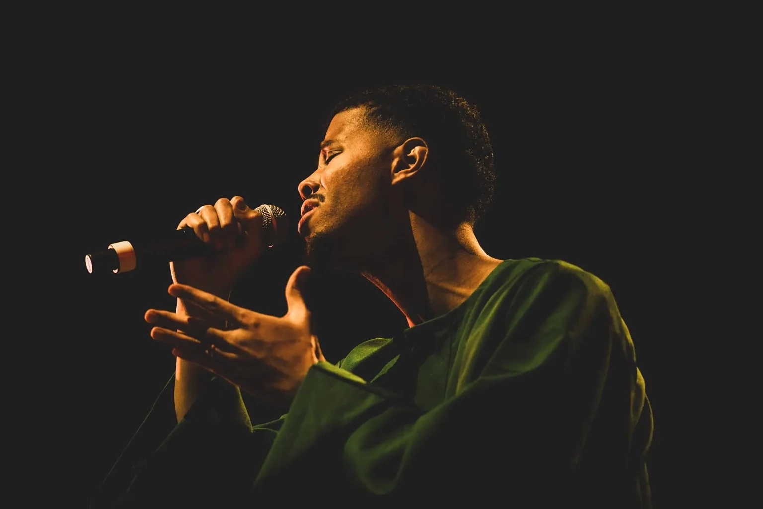 Mustafa sings onstage in a green shirt, holding a microphone against a dark background.