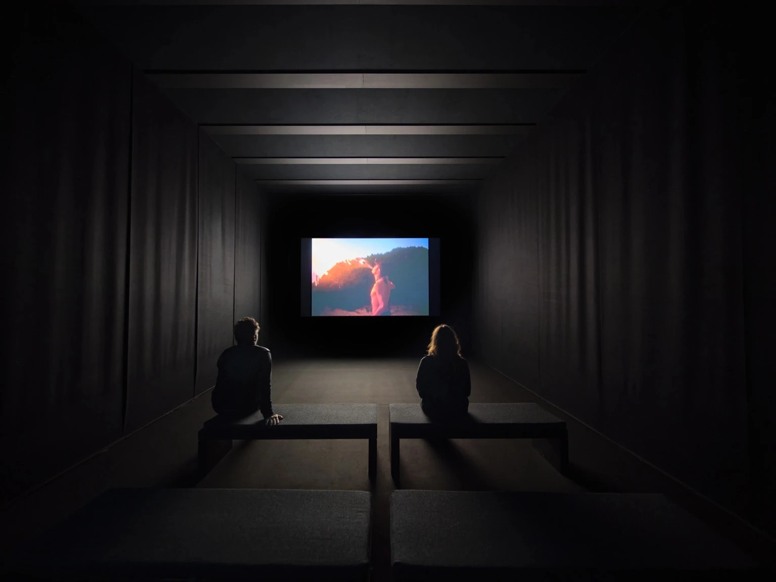 Two people sit on benches in a dark gallery watching a projected scene from Nan Goldins film Memory Lost at Pirelli HangarBicocca in Milan.