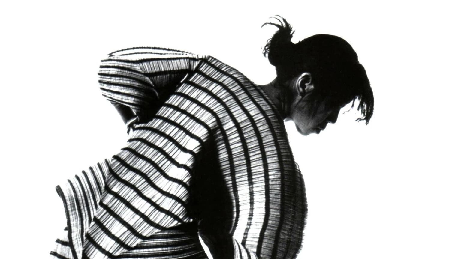 Black-and-white fashion photograph of a model in profile bending forward, wearing a sculptural striped pleated garment against a plain white background.