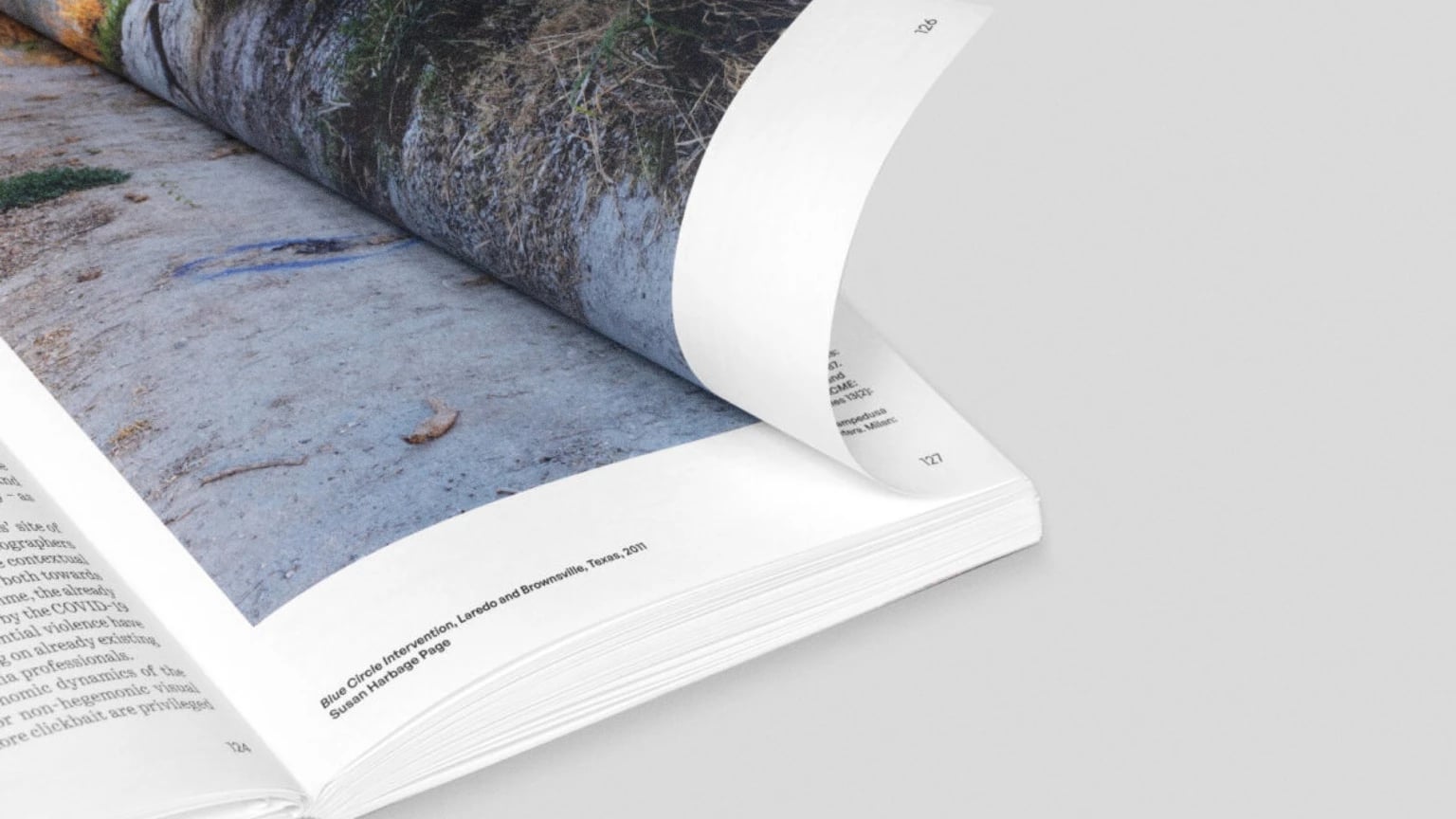 Close-up of an open photobook with a page curling over, revealing a landscape photograph printed across the spread on a light grey background.