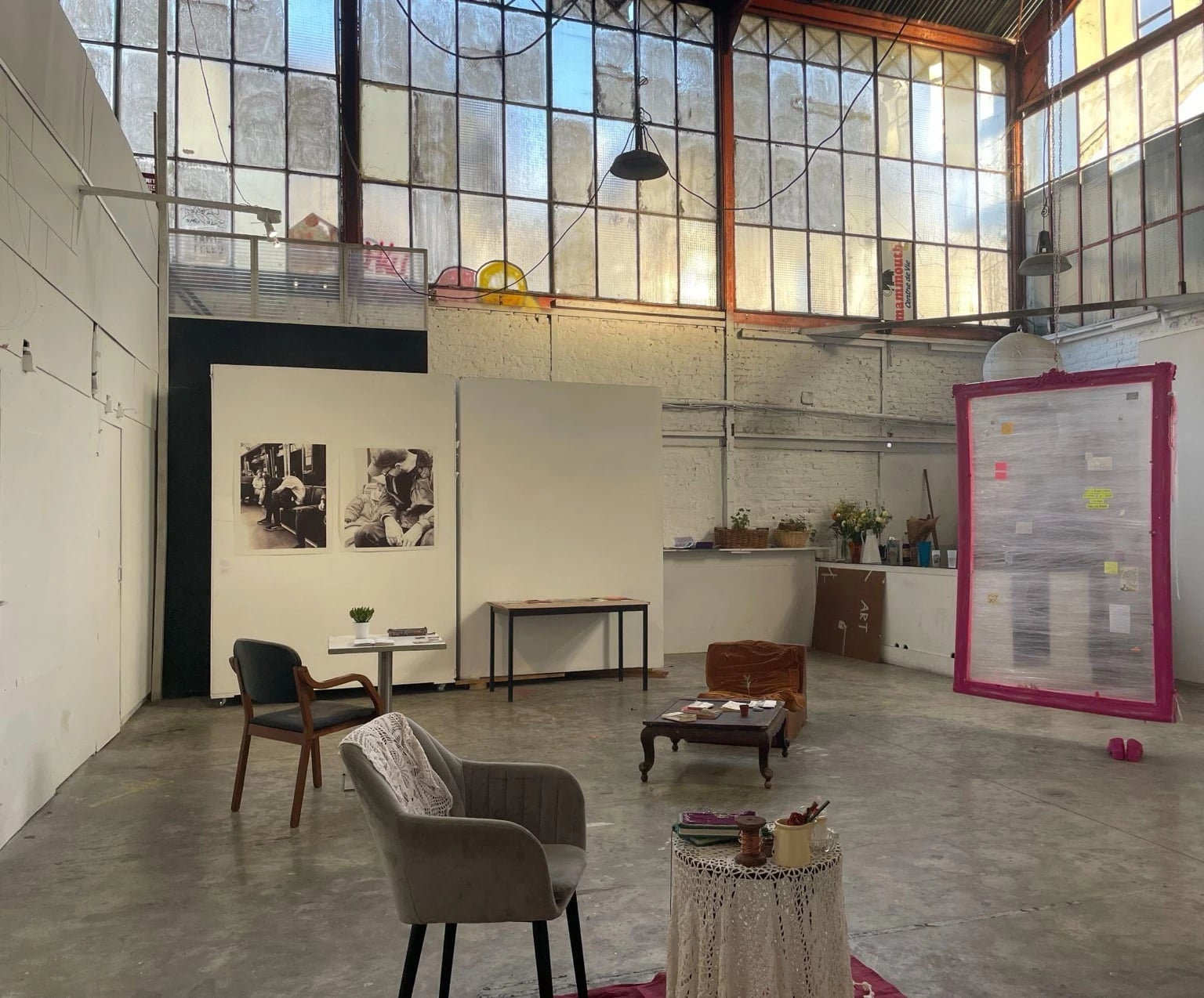 Bright industrial studio interior at Studio Albatros with high factory windows, minimalist furniture, photographic works on the wall, and a large framed installation wrapped in plastic.