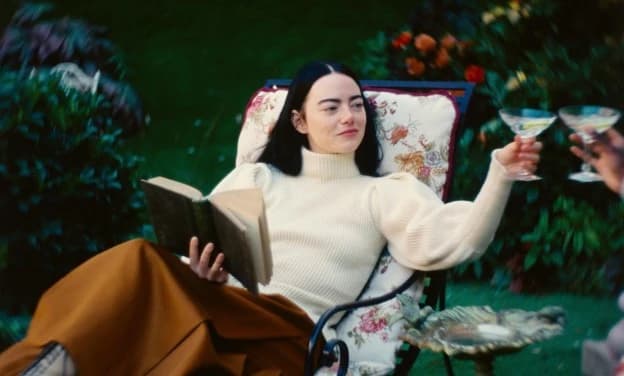 Emma Stones character lounges on a garden chair in Poor Things, holding a book in one hand and raising a cocktail glass in the other, surrounded by lush greenery and flowers.