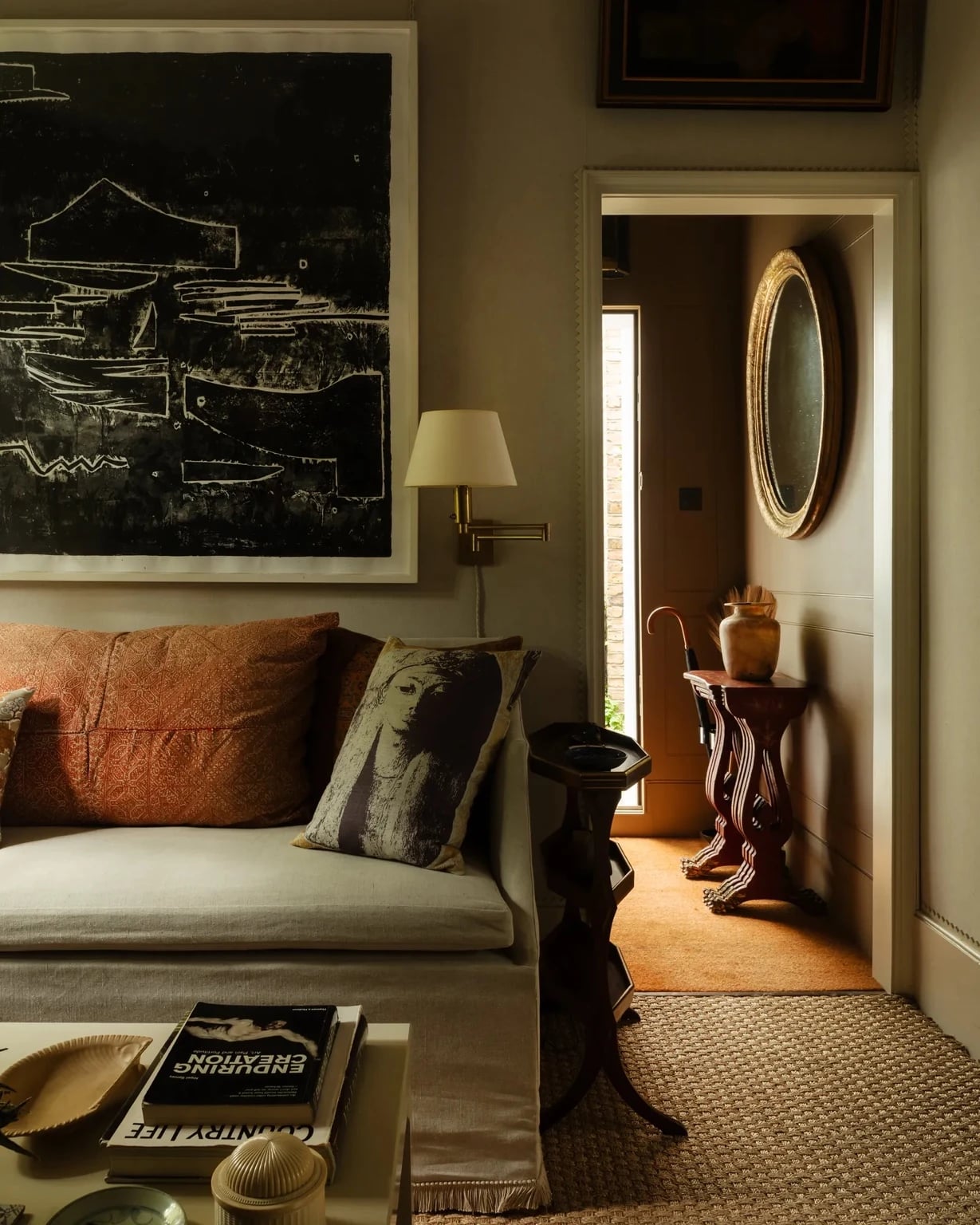 Warm living room with a cream sofa, patterned rust cushions, large black abstract artwork, and a view into a hallway with sculptural side tables and an oval mirror.