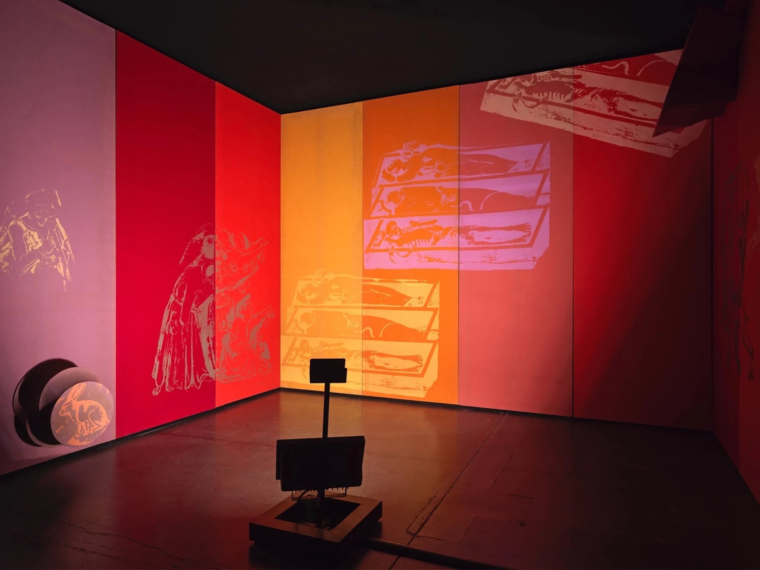 Installation view of Fabio Mauris work with large wall panels in gradients of pink, red, and orange, featuring faint graphic silhouettes of figures and coffins, with a dark floor and a small sculptural element in the foreground.