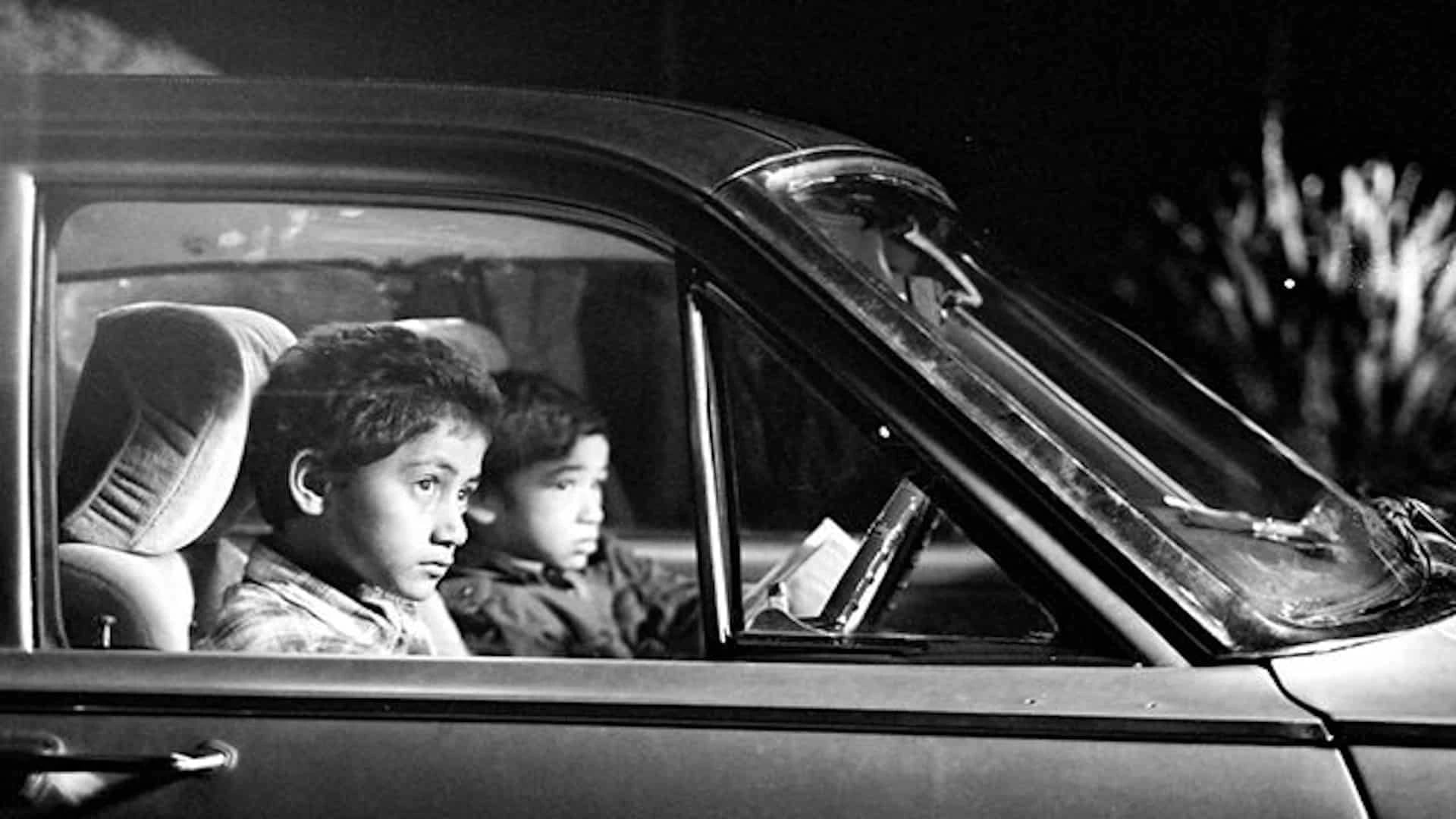 Black-and-white still from Two Cars, One Night (2003) showing two children sitting inside a parked car at night, captured in a quiet, intimate moment.