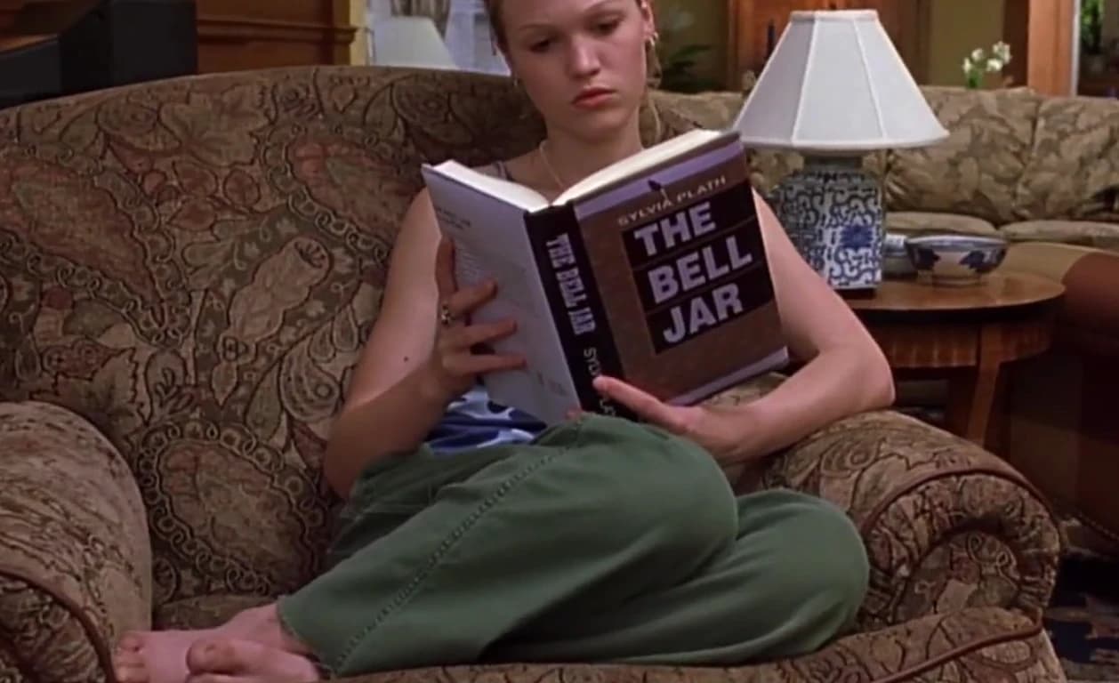 Film still of a young woman seated in an armchair reading The Bell Jar by Sylvia Plath, shown in a quiet domestic setting.