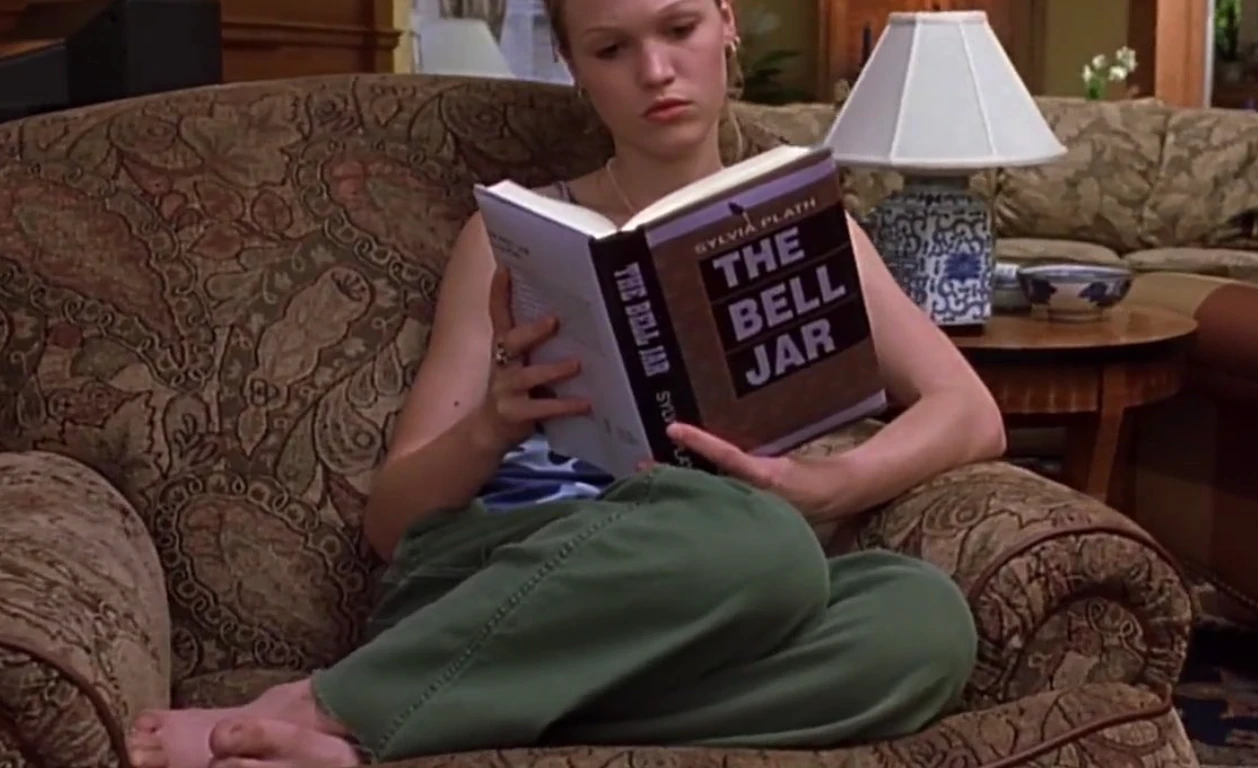 Film still of a young woman seated in an armchair reading The Bell Jar by Sylvia Plath, shown in a quiet domestic setting.