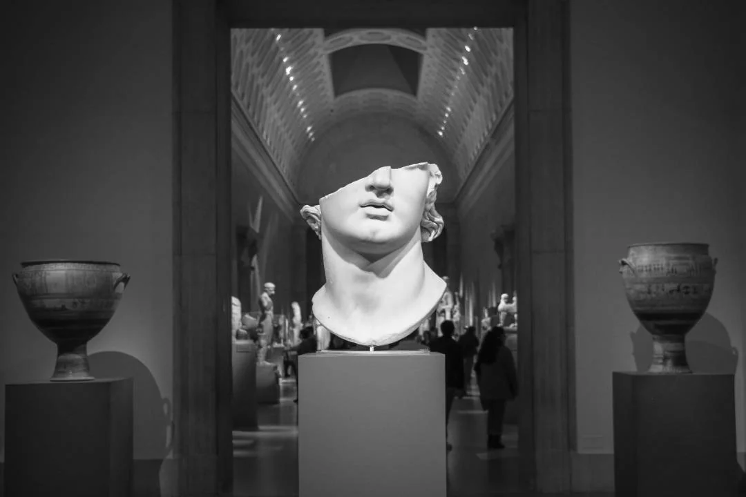 Black-and-white museum gallery view with a large classical marble bust on a pedestal framed by doorways, with visitors in the background.