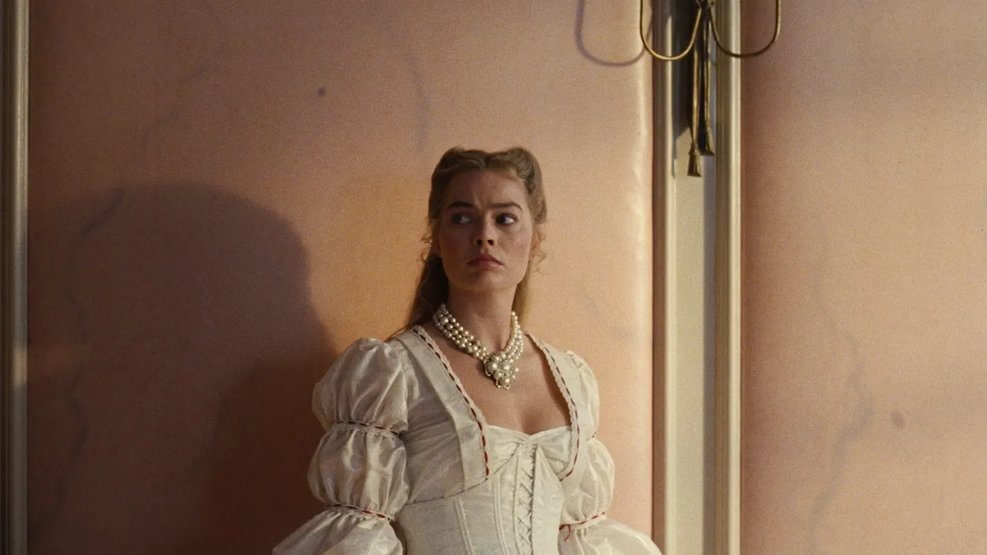 A cinematic still from Wuthering Heights featuring a woman portrayed by Margot Robbie standing in a windswept landscape, dressed in period costume, with soft natural light and muted tones creating a moody romantic atmosphere.