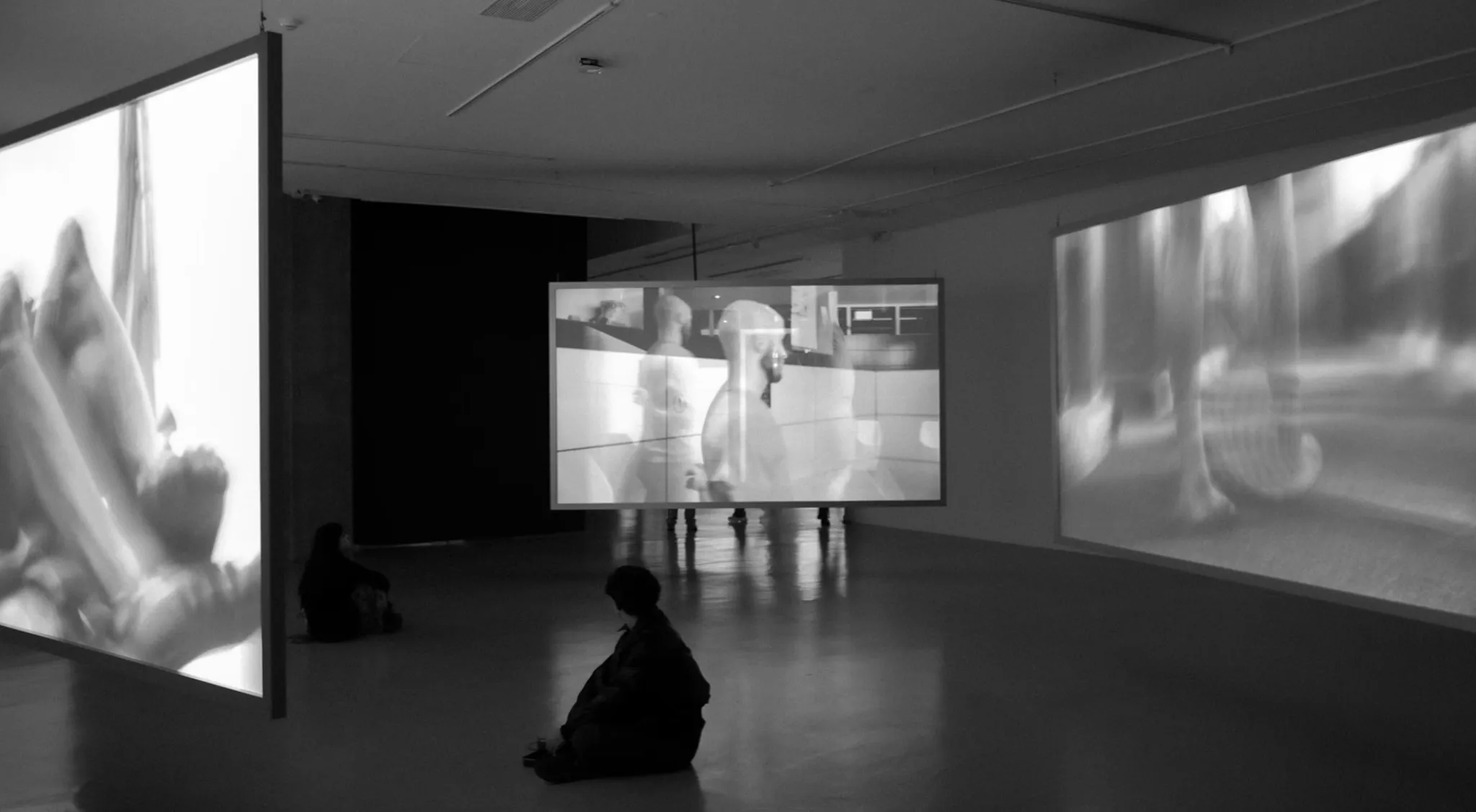 A dark gallery space with multiple large scale black and white video projections displayed on walls and a suspended screen, while visitors sit and watch the immersive installation