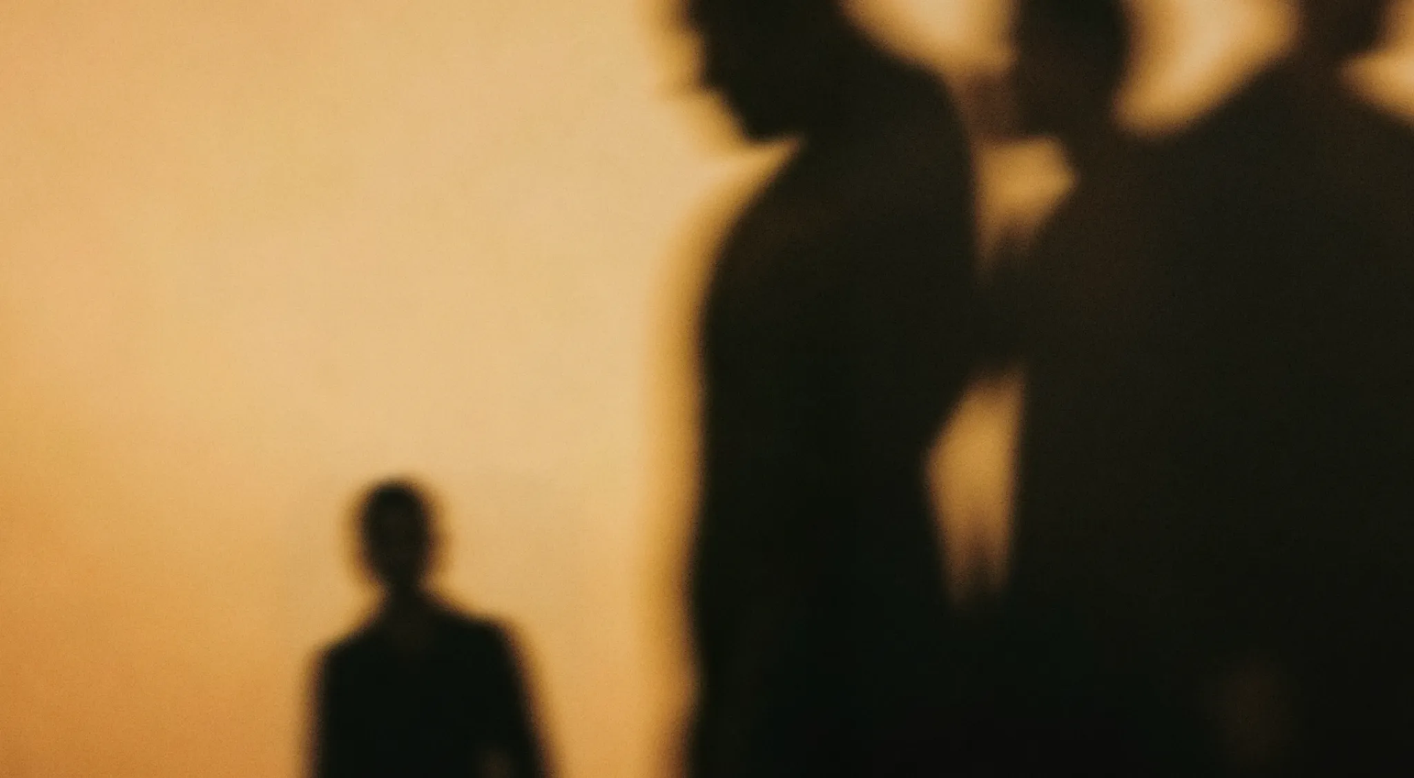Softly blurred silhouettes of several figures standing against a warm golden background, creating an abstract, cinematic scene with strong light and shadow