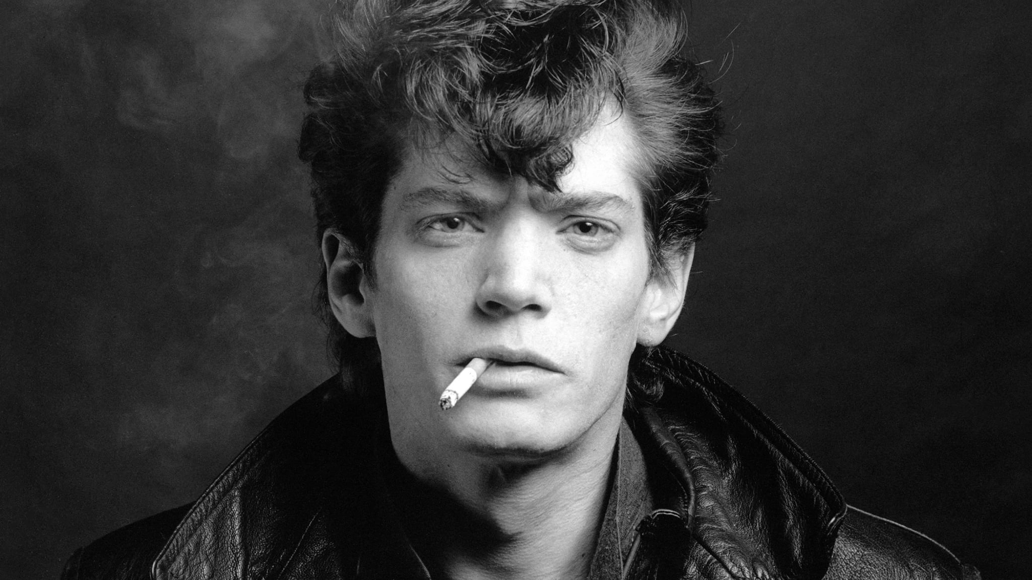Black and white portrait of Robert Mapplethorpe with tousled hair and a cigarette held between his lips, wearing a leather jacket and looking directly at the camera. Self Portrait, 1980 Robert Mapplethorpe Foundation