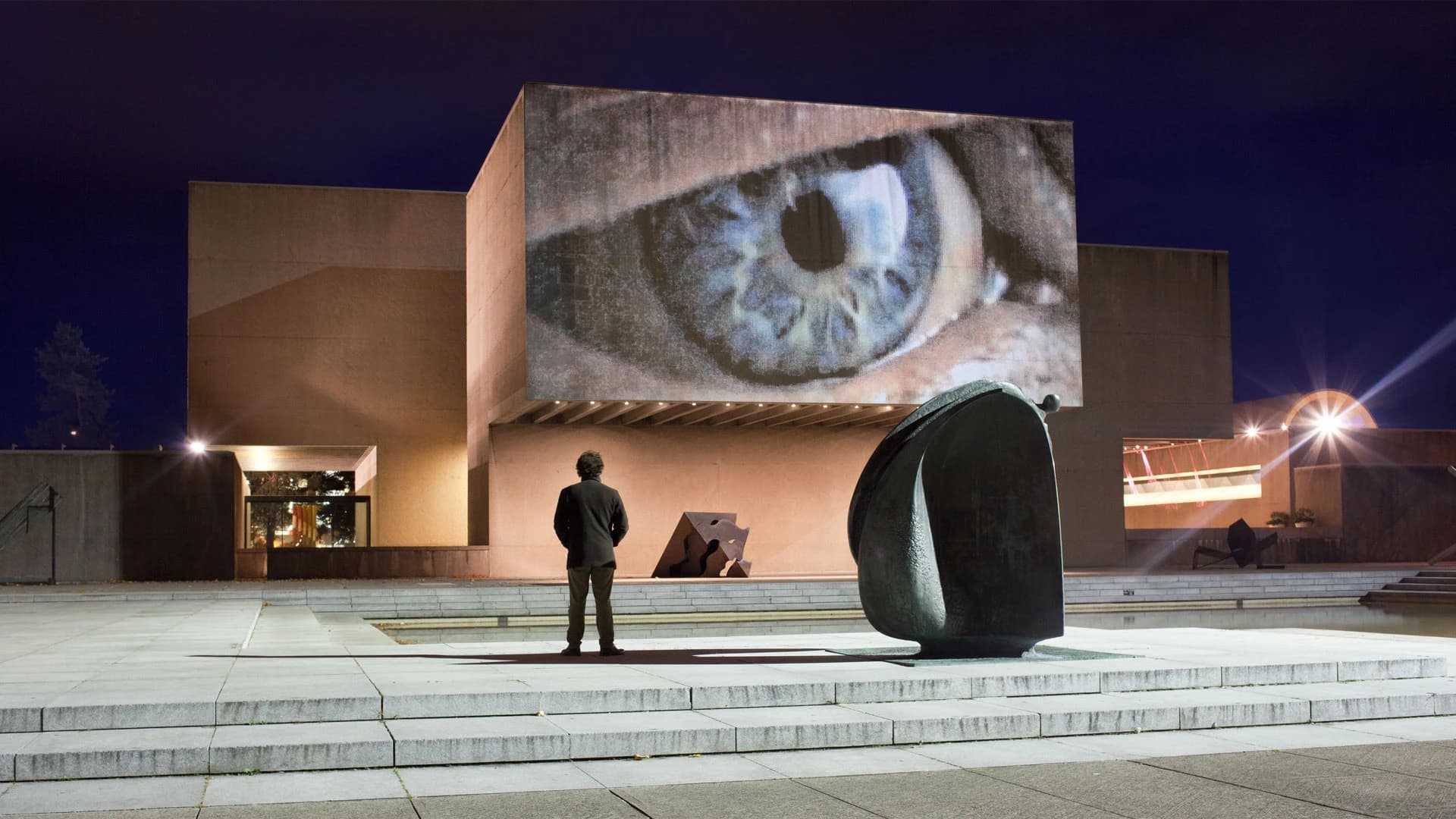 Night-time view of a public video installation by The Otolith Group showing a large projected close-up of an eye on a modernist building facade, with a single viewer standing in the plaza below.