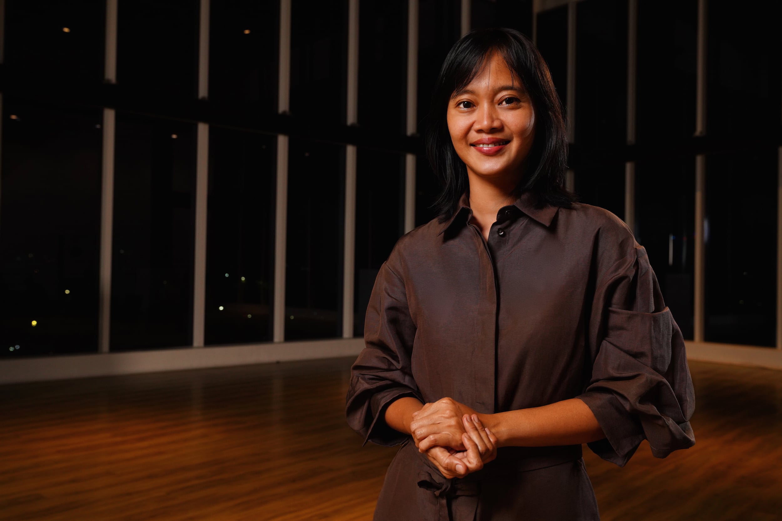 Dian Suci smiling in brown shirt standing in modern interior at night