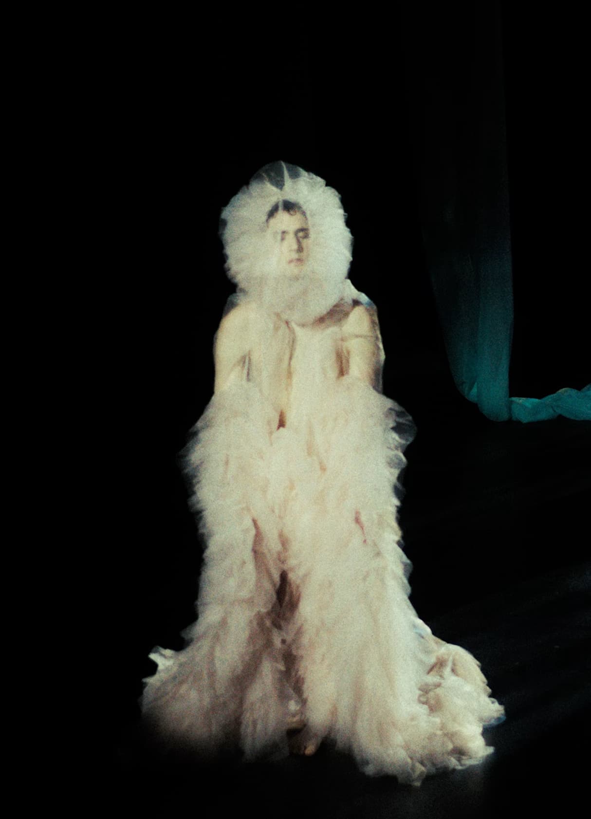 Model wearing voluminous white tulle gown with ruffled halo headpiece on dark stage