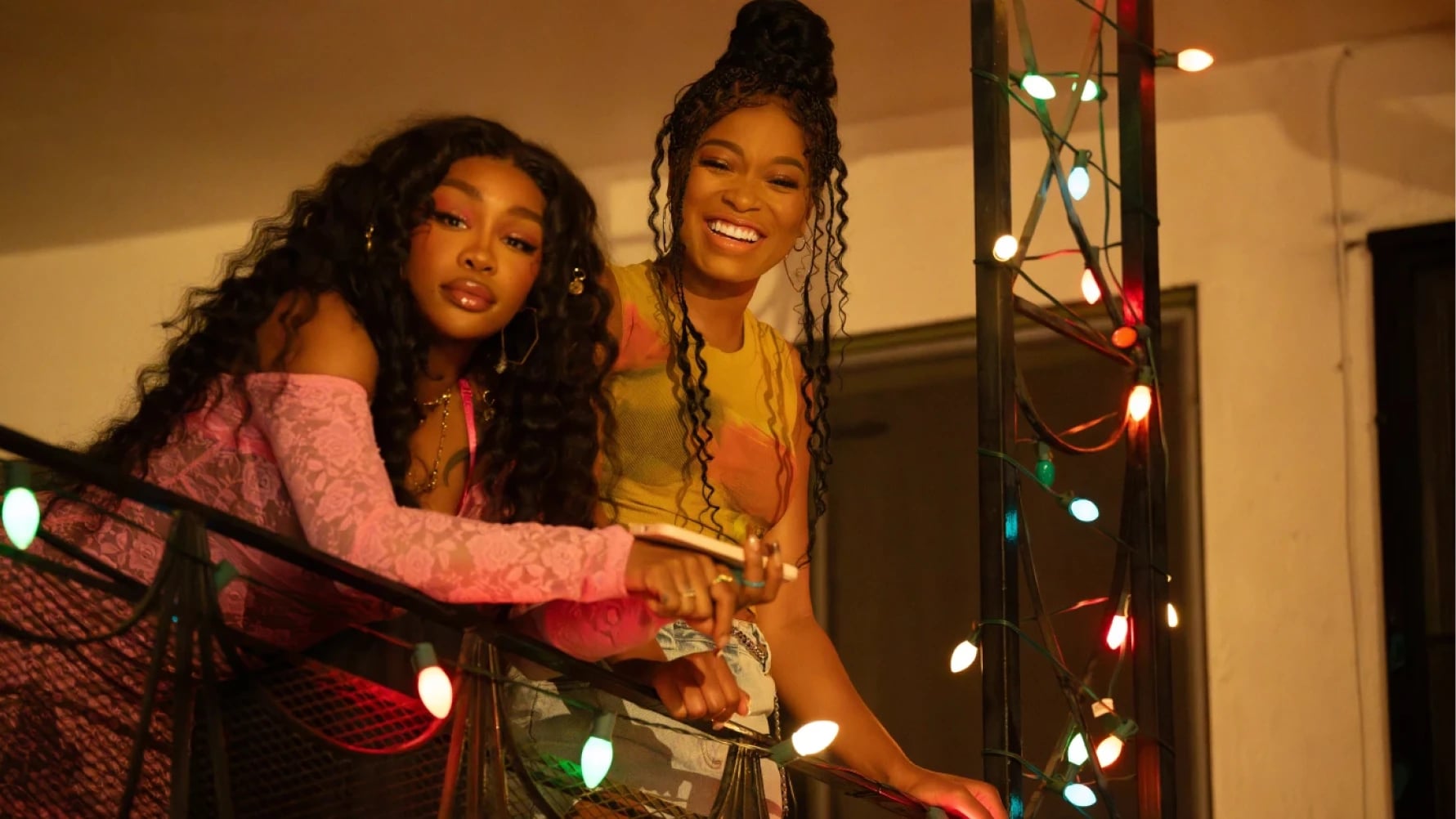 Two women smiling on a festive light-decorated balcony in a scene from One of Them Days 2025