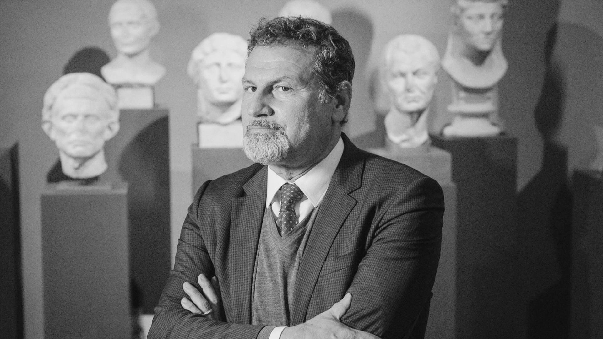 Massimo Osanna stands arms crossed among classical marble busts in a black-and-white portrait