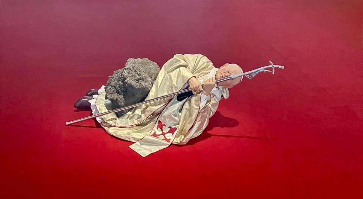 Maurizio Cattelan’s true to size multi media sculpture showing Pope John Paul II stuck by a meteorite lying on a deep red moquette-like floor.