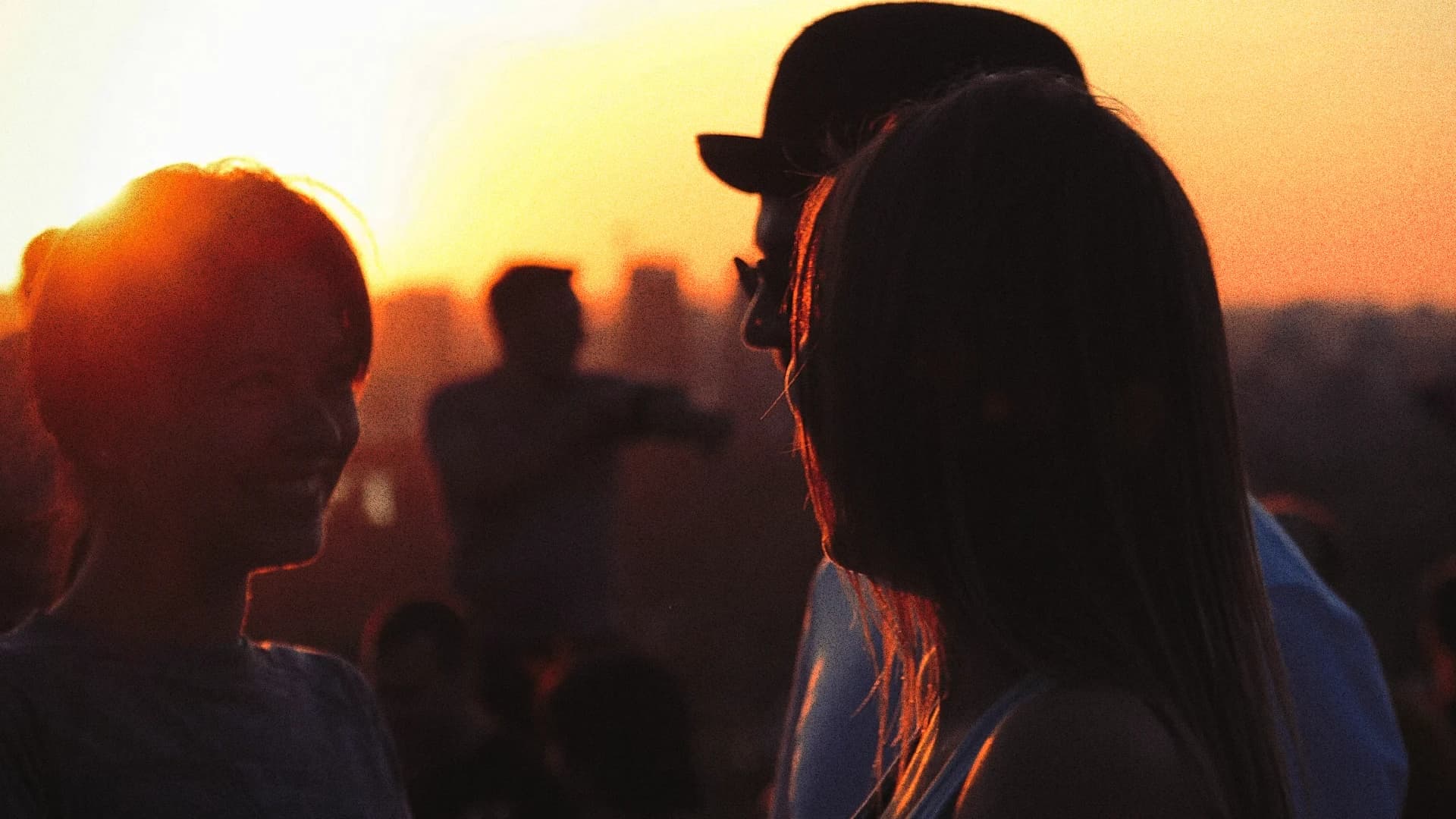 Two people in silhouette at a golden hour outdoor gathering, warm sunset glow and crowd in background