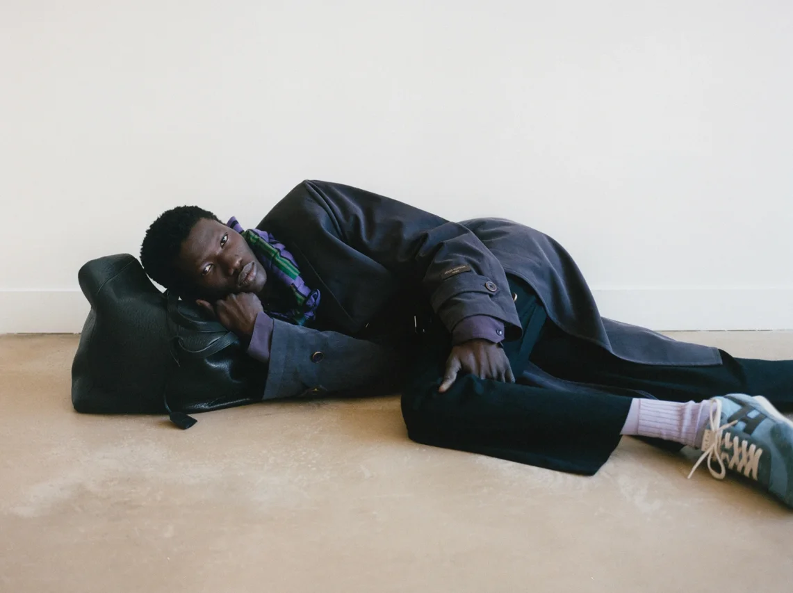 Fashion photograph of a man lying on the floor with his head resting on a black leather bag, wearing a layered outfit of a dark brown coat, purple and green plaid shirt, gray trousers, pink socks, and sneakers, against a plain white wall and beige floor.