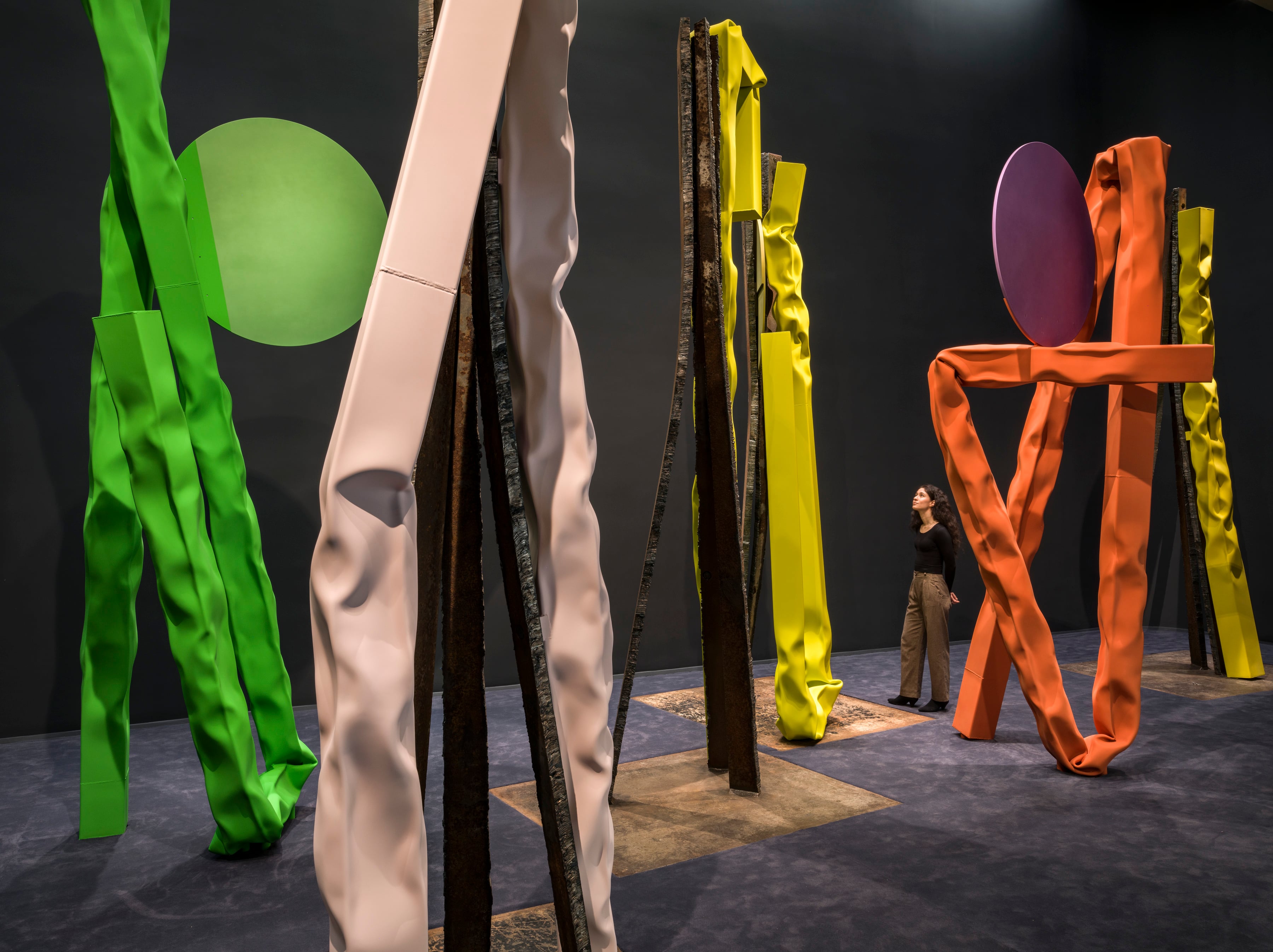 Gallery installation view of large sculptural works by Carol Bove, featuring tall twisted fabric-like forms in green, beige, yellow, and orange draped over rusted steel beams, with a purple oval disc and a figure viewing the artworks in a dark exhibition space with a stone-toned floor.