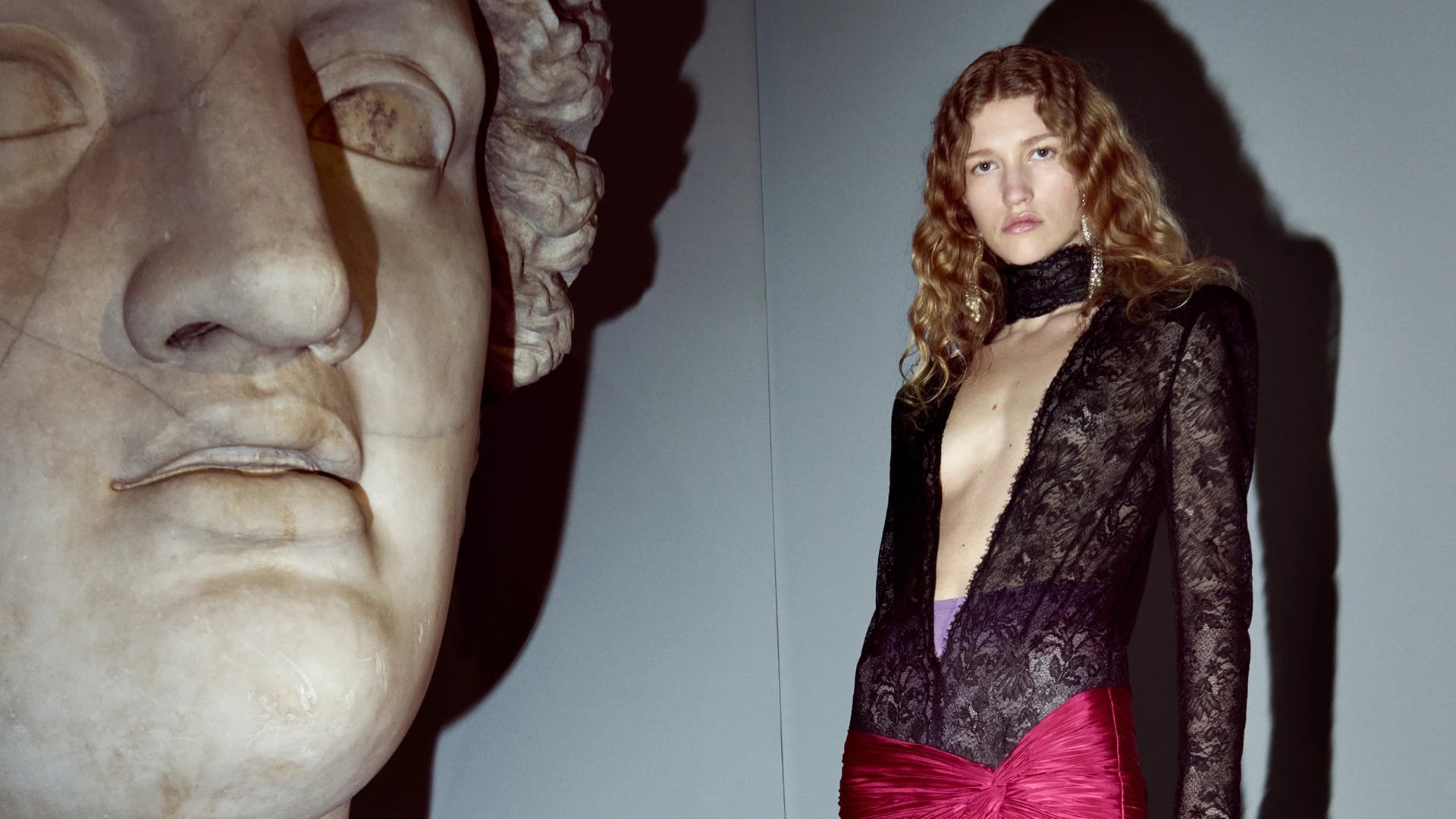 Valentino Interferenze FW26 campaign: model in black lace bodysuit with magenta satin skirt beside marble sculpture