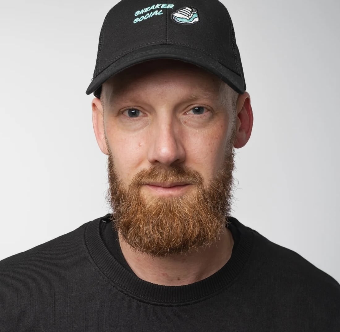 A bearded man faces the camera with a neutral expression, with a black cap atop his head featuring words and an image in blue.