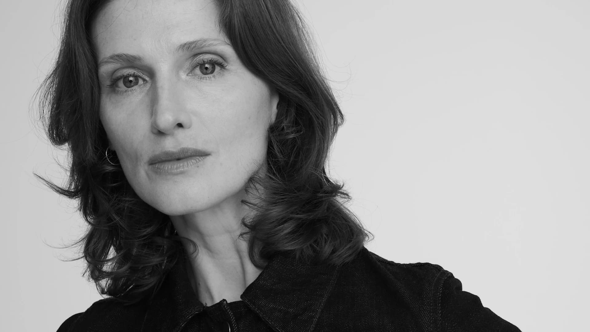 Black-and-white studio portrait of a woman with wavy shoulder-length dark hair, wearing a black denim jacket, standing with hands on her hips, gazing directly at the camera against a plain light background.