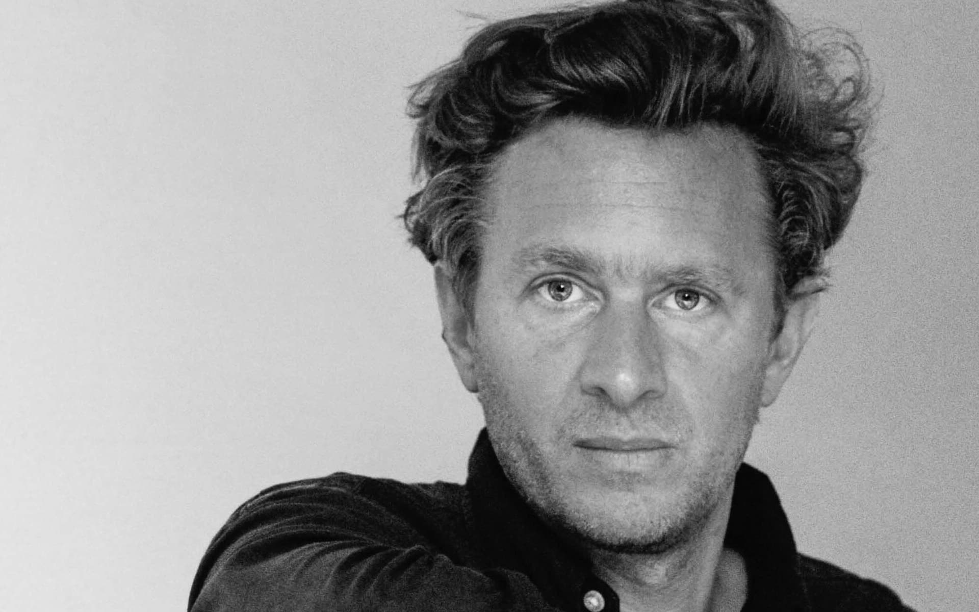 Black-and-white portrait of a man with wavy dark hair, wearing a dark shirt, leaning his arm on a tall wooden panel while gazing directly at the camera against a plain light background. Nicolas Schuybroek