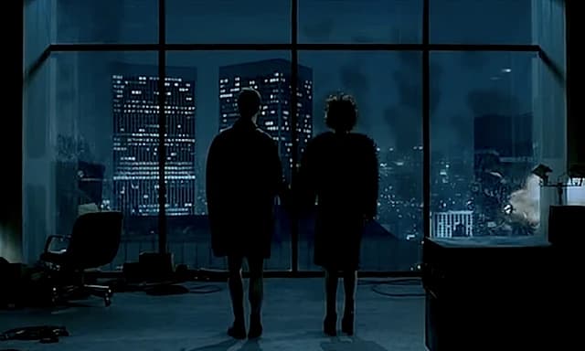 Silhouetted couple standing hand in hand before a wall of windows overlooking a nighttime city skyline, with buildings visibly exploding and collapsing in the background, from the final scene of "Fight Club."