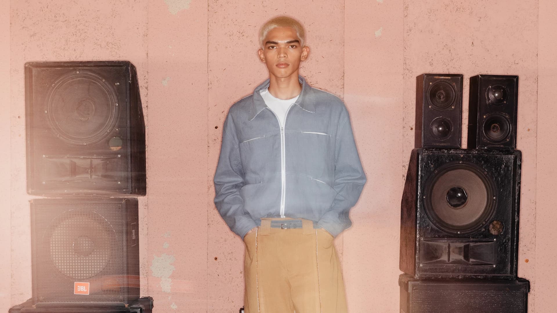 BAGASAO menswear look featuring light blue zip jacket and tan trousers styled against pink wall with JBL speakers